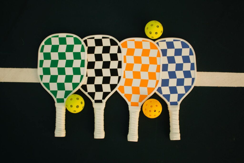 Four colorful pickleball paddles in green, black, orange, and blue, arranged alongside a yellow pickleball on a court backdrop.