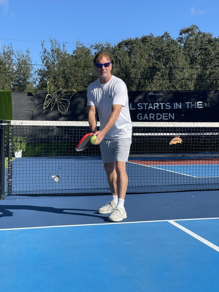 A man stands on a blue tennis court, preparing to hit a tennis ball with a racket, under a clear sky. A sign in the background reads, "All Starts in the Garden."